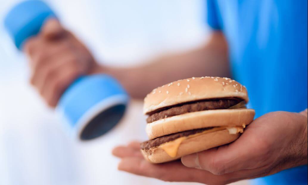 Homem com um peso em uma mão e um hamburguer em outra